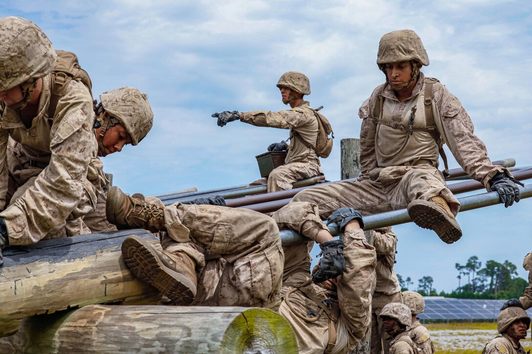 Marine Corps recruits climb above and under slanted bars under a cloudy sky.