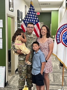 U.S. Army Soldier in uniform poses with wife, son and daughter in front of an American Flag and a USAREC Flag.