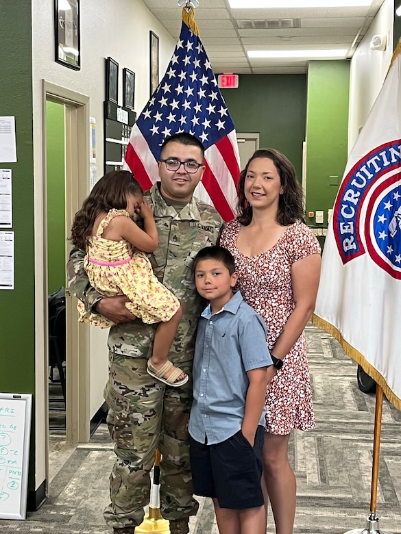 U.S. Army Soldier in uniform poses with wife, son and daughter in front of an American Flag and a USAREC Flag.