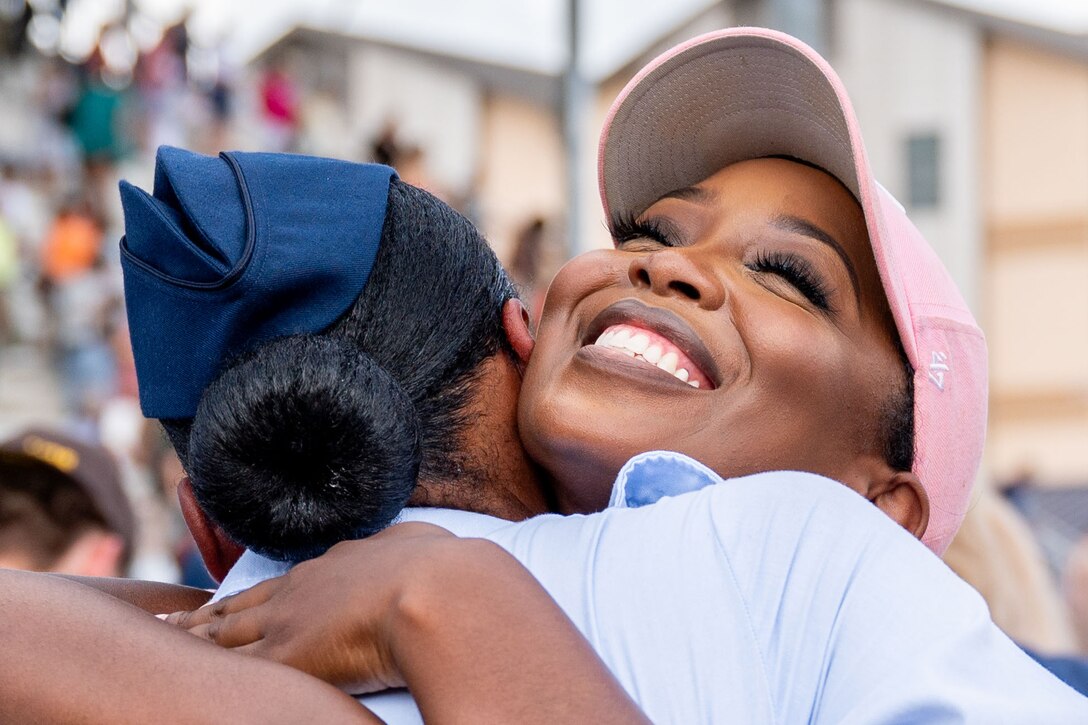 A close-up of a family member hugging an Air Force trainee with people standing in bleachers next to a building in the blurred background.