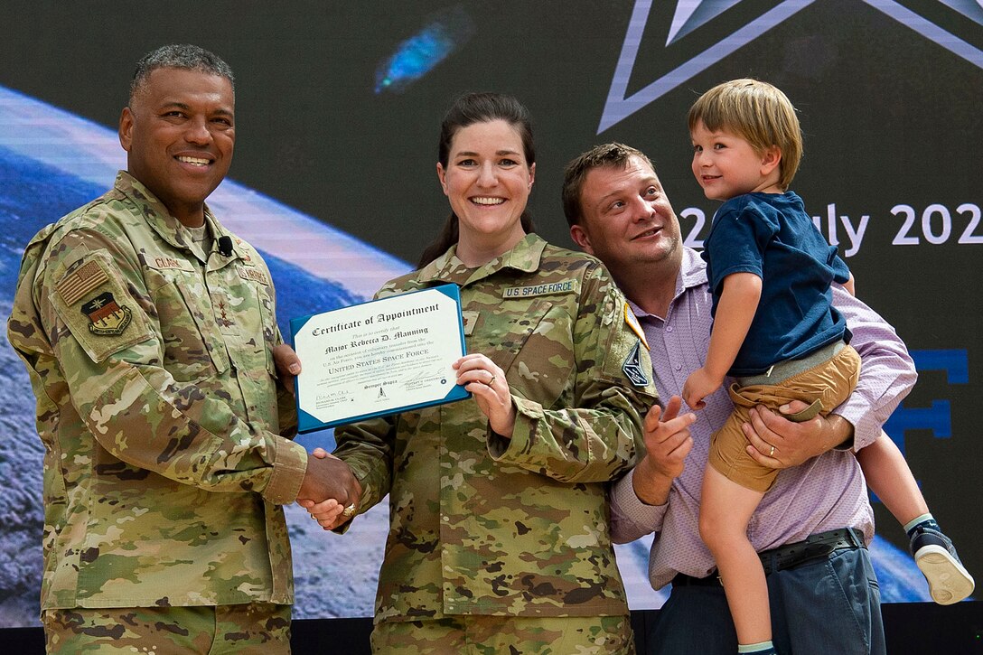 A guardian shakes hands and jointly holds up a certificate with an airman while standing on a stage with a civilian adult holding a child.