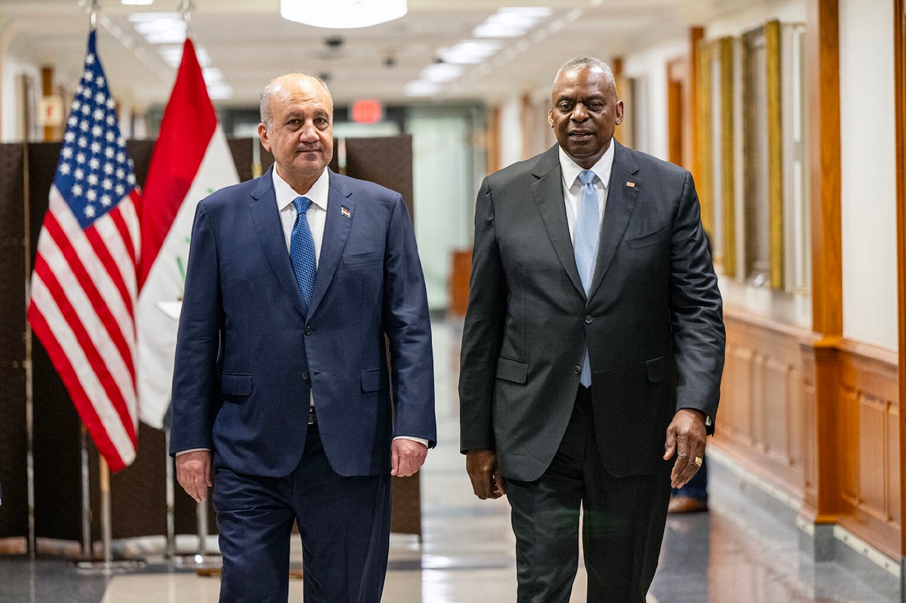 Two people in business attire walk down a hallway with flags behind them.