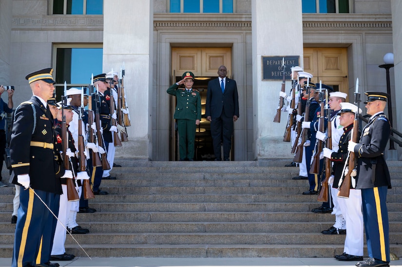 A man in a foreign military uniform salutes while standing next to a man in business attire.