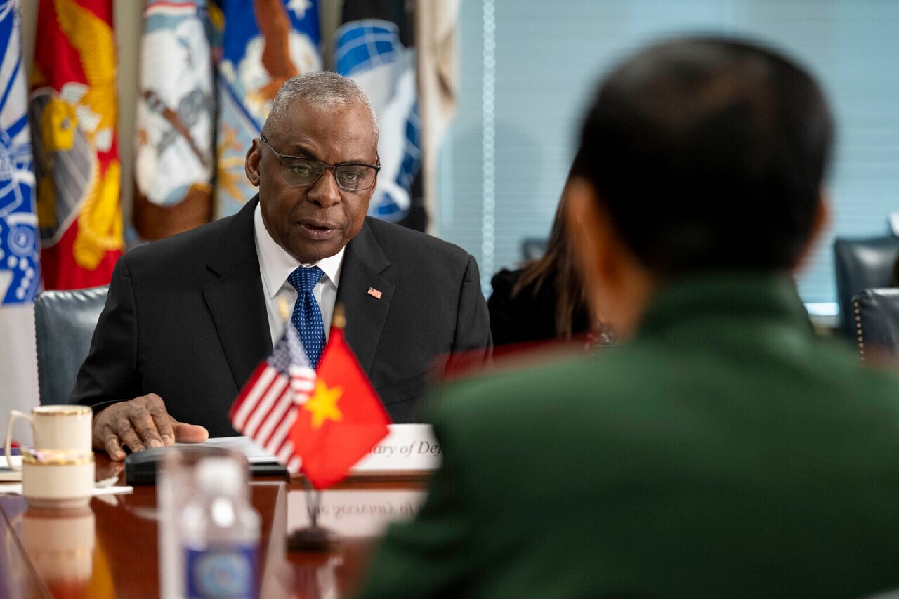 A man in business attire sits at a table displaying U.S. and Vietnamese flags.