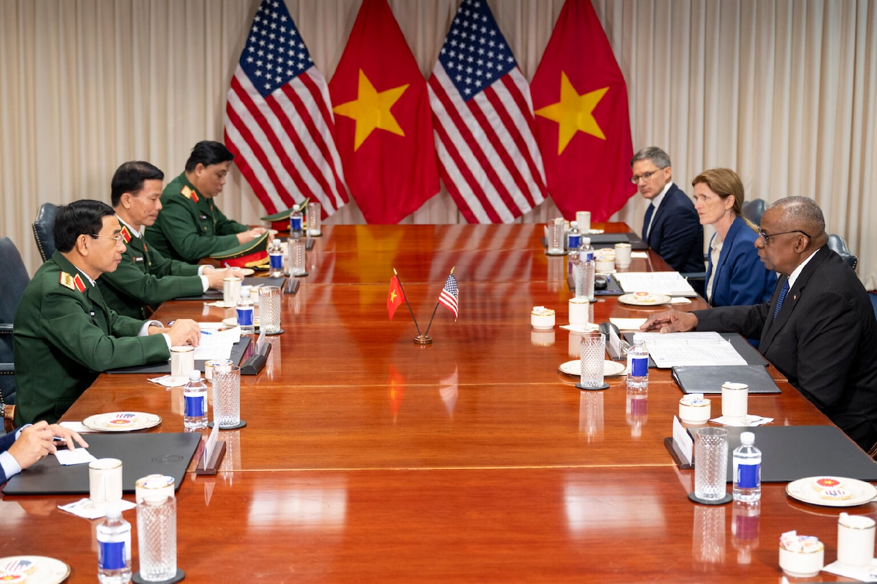 A group of men in foreign military uniforms sit at a table across from men and women in business attire.