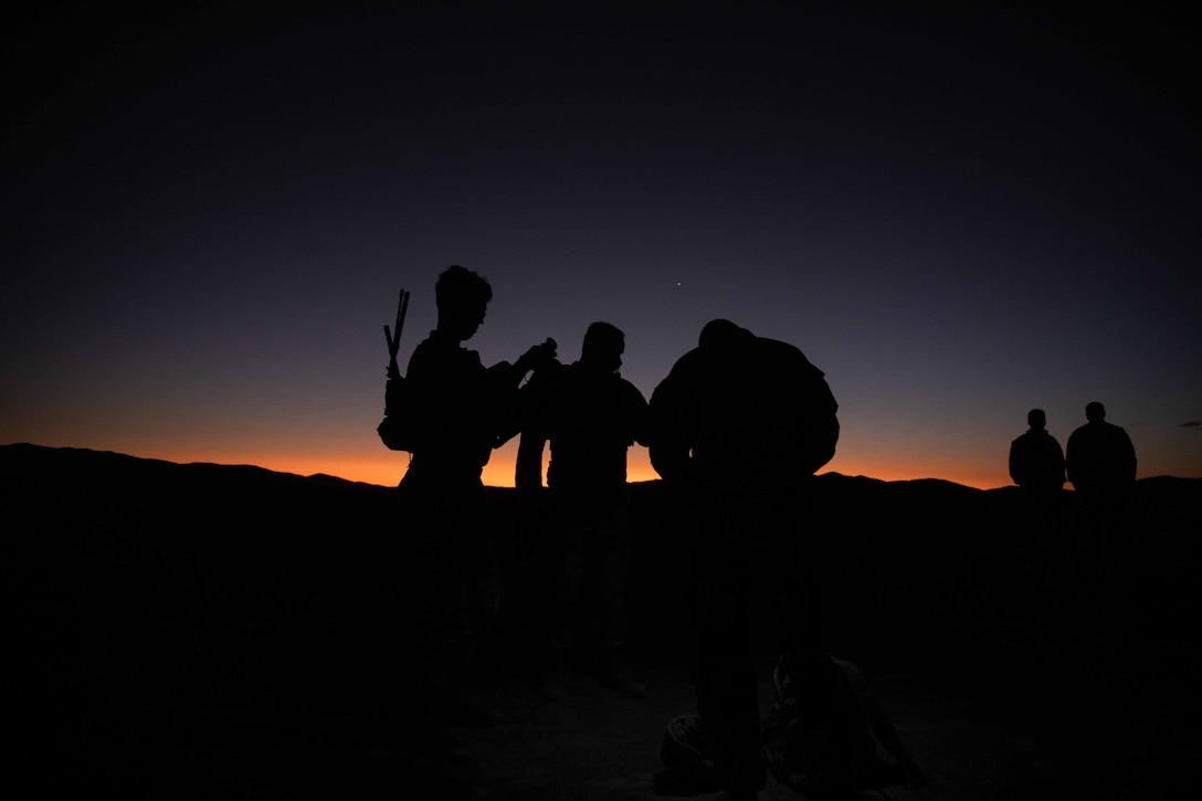 Soldiers are shown in silhouette against an orange light on the horizon as they set up equipment in an open area.