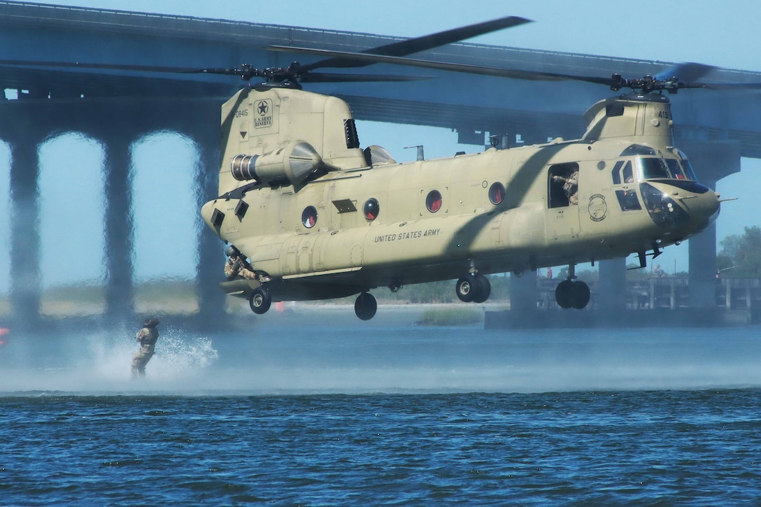 A soldier conducts a daytime jump from a low-flying helicopter into a river. A large bridge is in the background.