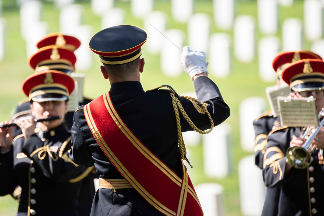 A soldier seen from behind conducts a small band in formal uniforms against the backdrop of a cemetery.