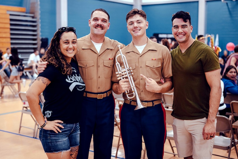 Two Marines, one holding a musical instrument, are flanked by civilians inside a hall with people in the background.