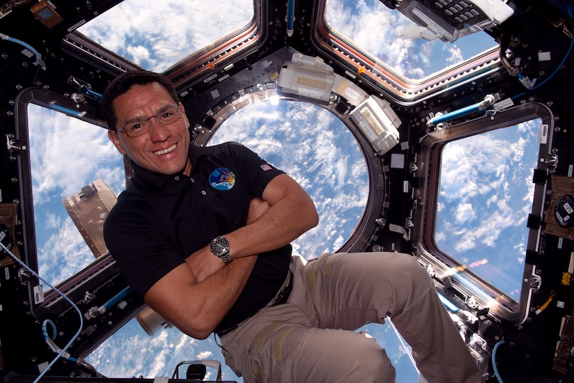 A man wearing business casual attire poses for a photo with his arms crossed while floating aboard the International Space Station; the Earth is visible through windows in the background.