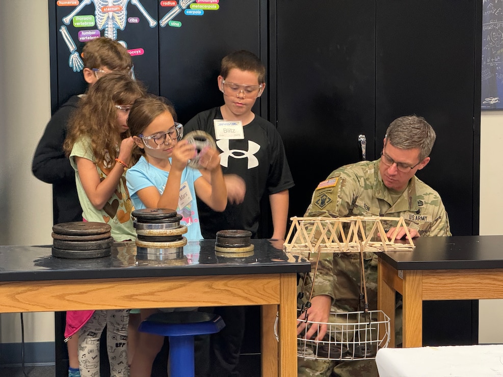 A man in uniform, adds weights to a basket as four students eagerly watch.