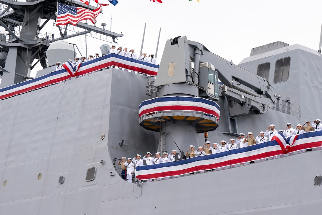 Sailors salute while standing on the decks of a ship on an overcast day.