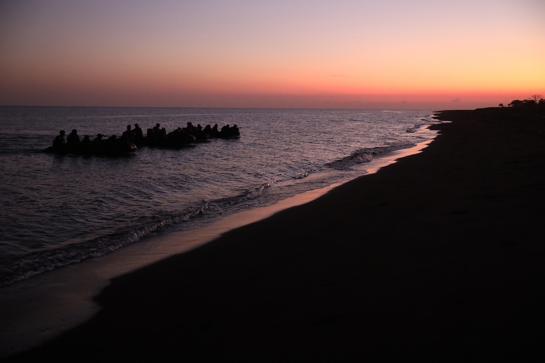 Service members in silhouette approach a shore in amphibious assault vessels at twilight.