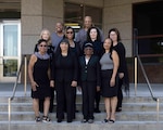 Ten women dressed in black dresses smile at the camera. They are standing on concrete steps outside in front of a large building.