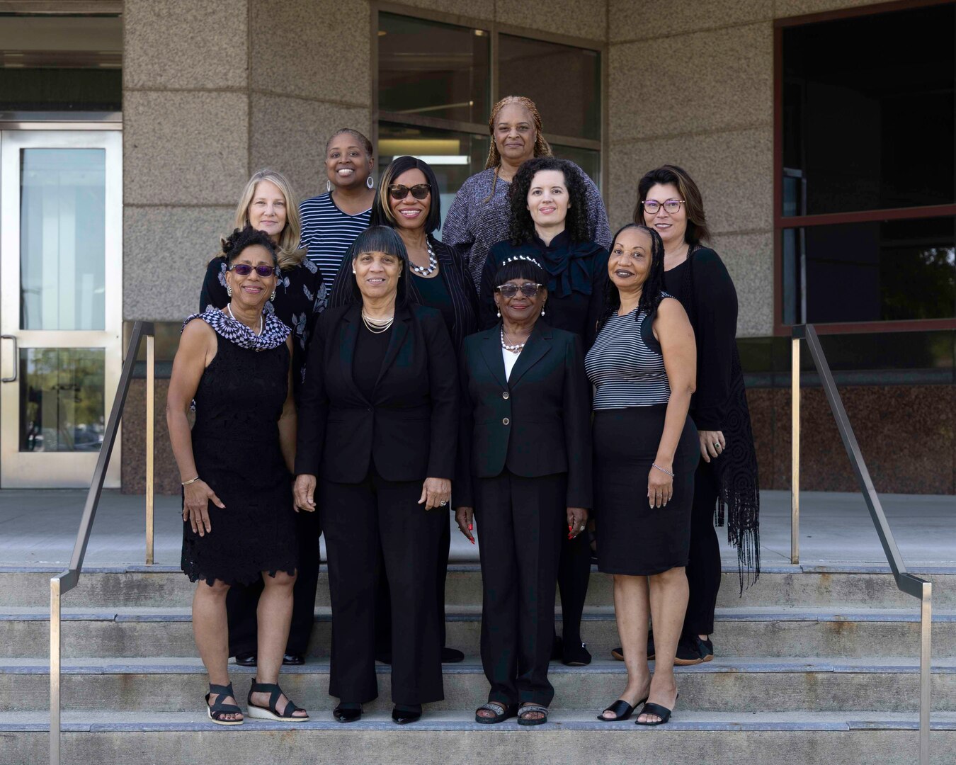 Ten women dressed in black dresses smile at the camera. They are standing on concrete steps outside in front of a large building.