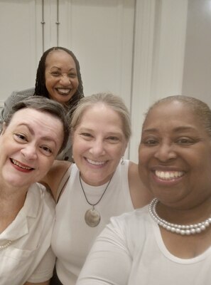 Four women, three dressed in white and one in a checkered blazer take a selfie pose and smile at the camera. Two are light skinned, two are dark skinned.