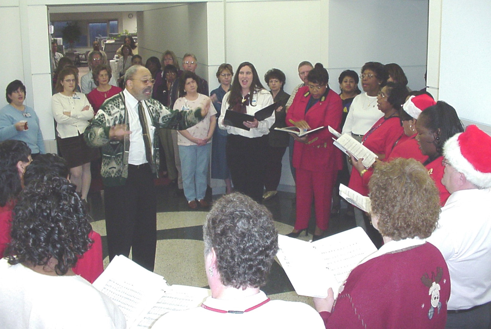 A group of people in holiday dress hold books and sing in a hallway. They are directed by a dark skinned man with graying hair.