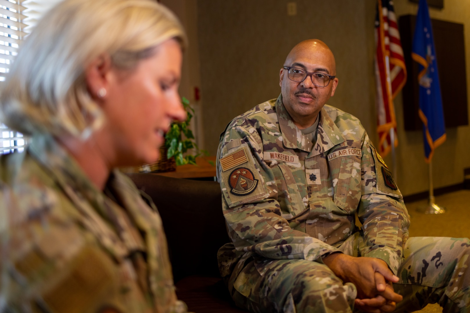 Uniformed service member sits and looks down; uniformed chaplain wearing glasses sits across from her.