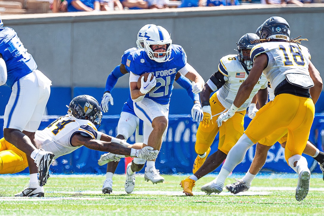 A football player runs with the football in between an opening of opposing players as one dives to grab their leg.