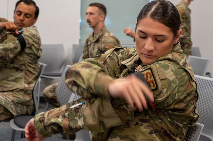A woman holds out her left arm and applies a tourniquet using her right arm. Others in the background do the same