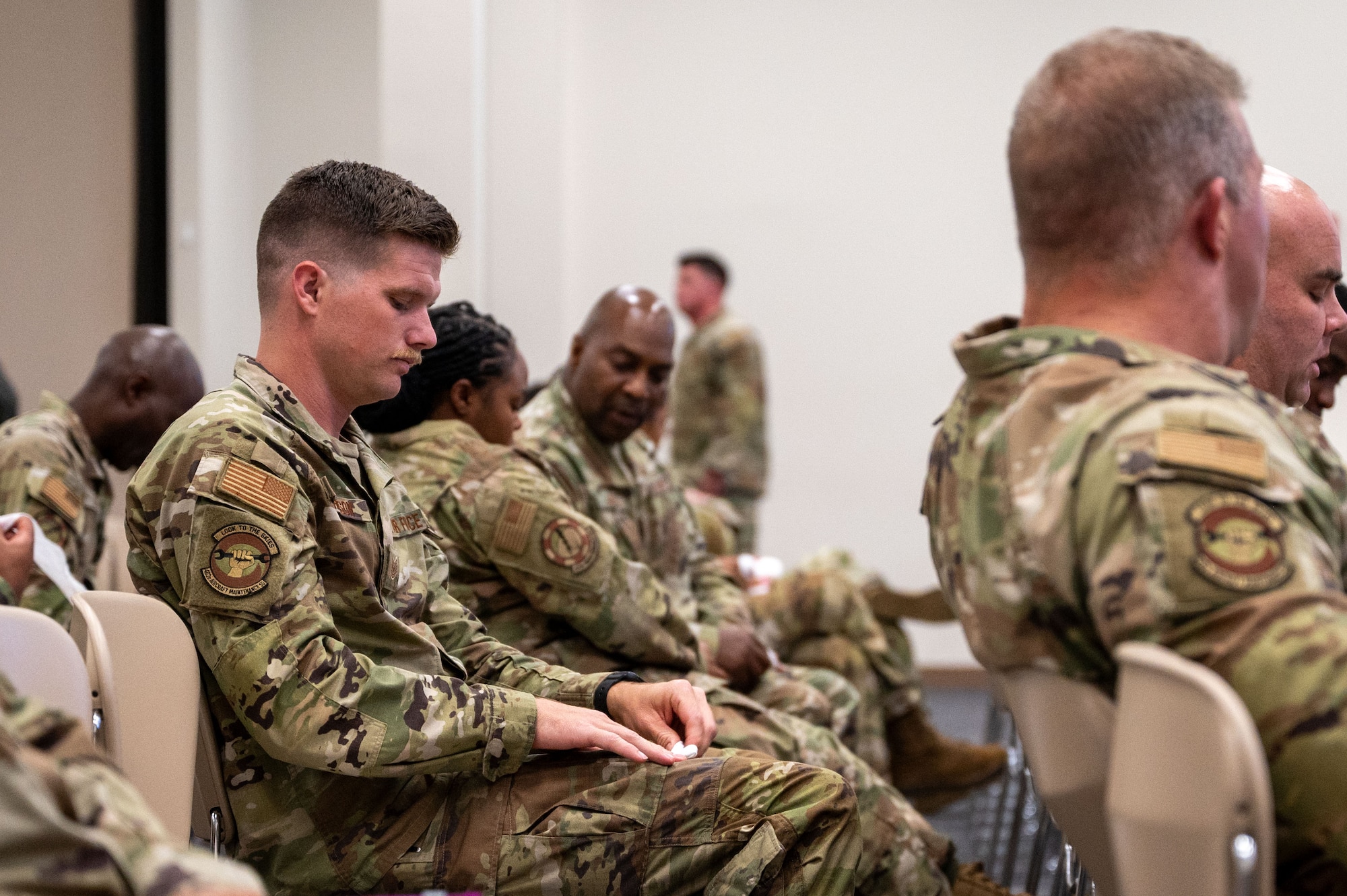 An Airman, seated among others, uses both hands to simulate applying a hemostatic dressing to a wound on his right thigh