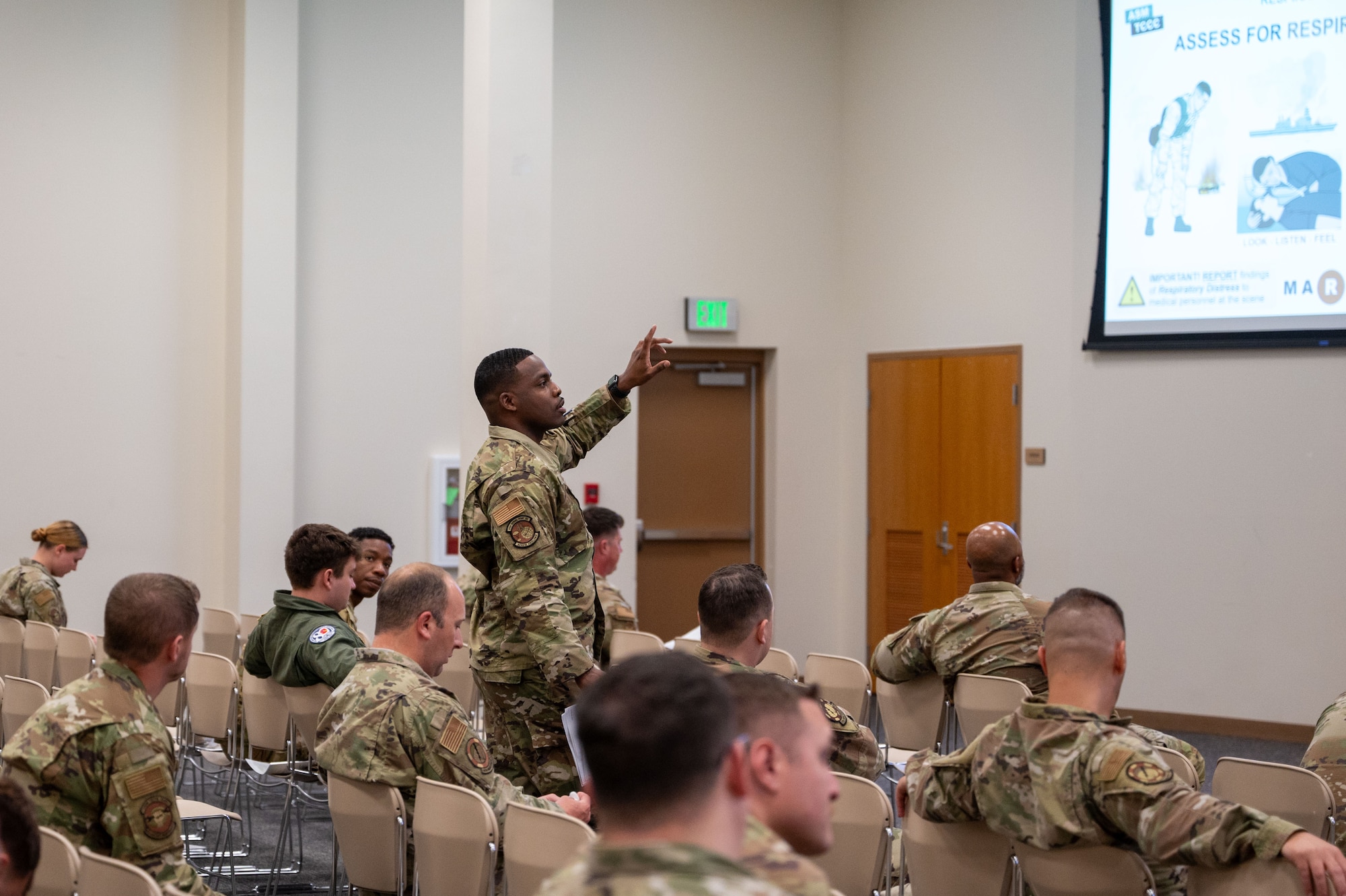 Among seated Airmen, Hamton stands in a row down the middle of an auditorium and points to a screen on the wall while talking