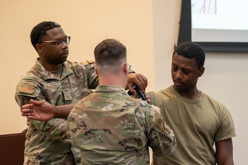 One man stands with his arm stretched out while two others stand on each side of his arm adjusting a tourniquet