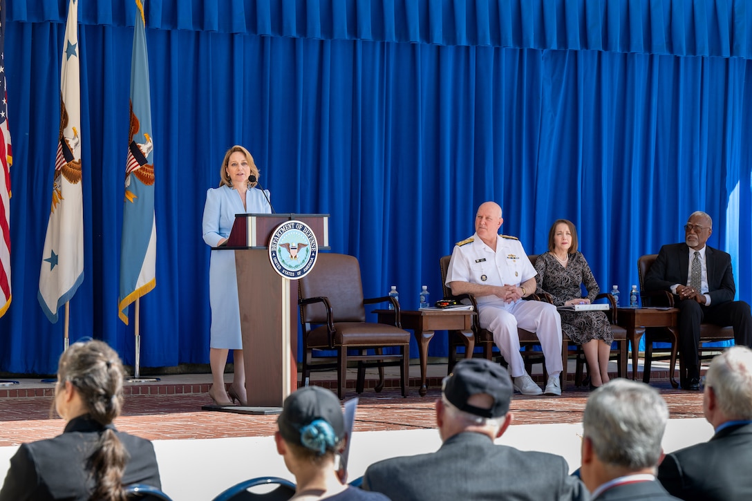 Deputy Defense Secretary Kathleen Hicks addresses a seated audience from a lectern on an outdoor stage where others sit.