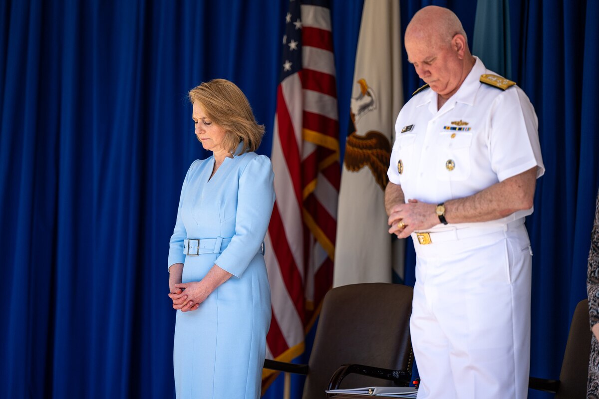 Deputy Defense Secretary Kathleen Hicks and Navy Adm. Christopher W. Grady bow their heads while standing in front of a blue curtain.