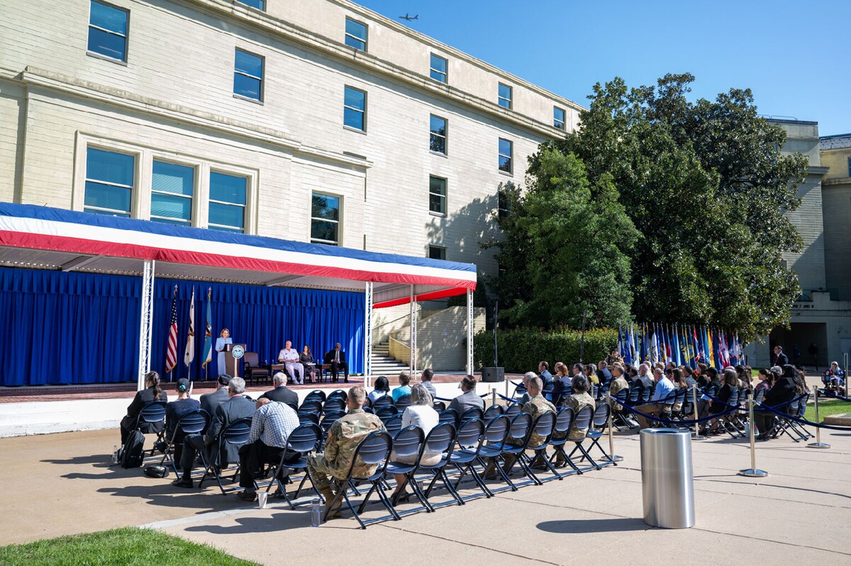 People are seated in rows of chairs outside of a large building, looking at a stage where a person is at a lectern.
