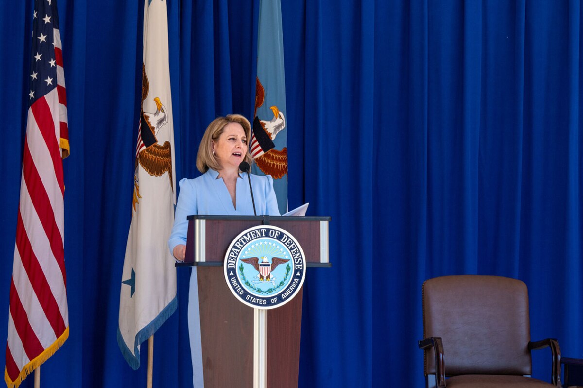 A person in business attire stands at a lectern delivering remarks to people off screen.