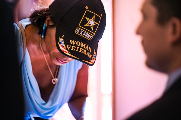 a woman leans over signing something, leaving only her u.s. army, woman veteran hat and lower jaw dramatically visible