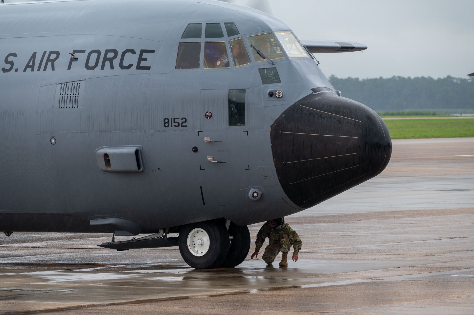 A c-130j sits on the flight line, a maintainer squats below the nose to check something