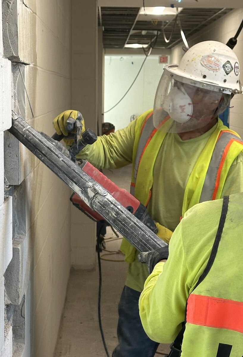 One worker operates a saw and cuts cement wall while other operates vacuum to remove dust.