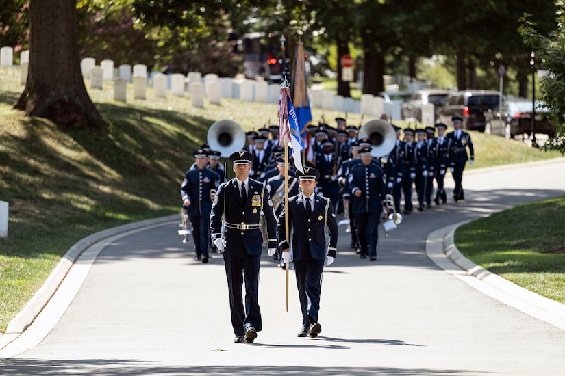 A group of airmen in formal attire parade on a road in a cemetery.