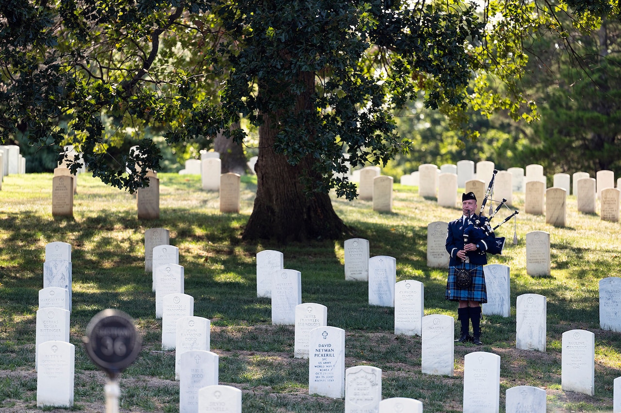 A bagpipe player stands in a field by a tree amid white tombstones.