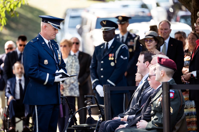 An airman stands in front of the deceased's family with an open booklet while others gather around.