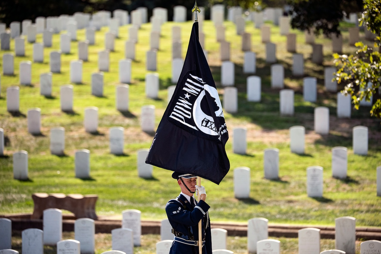 An airman holds the POW-MIA flag straight up while standing in the middle of many white tombs at a cemetery.