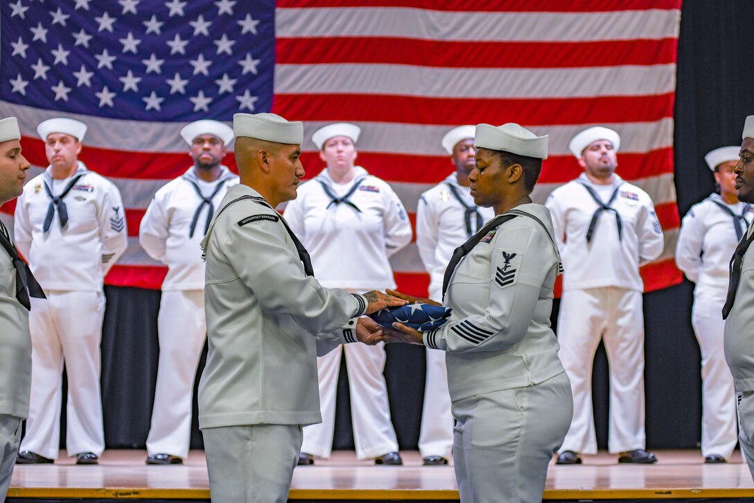 Two sailors pass a folded American flag as fellow sailors stand in formation behind them in front of a large American flag.