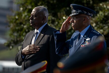 A person in business attire holds their hand over their heart as a second person in a military uniform salutes during a ceremony.