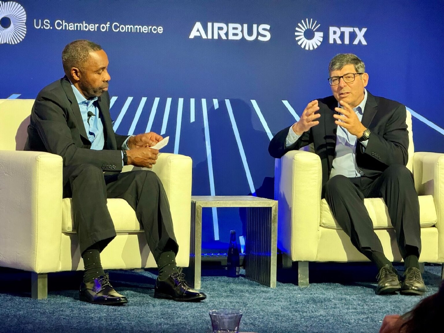 Two men are seated next to each other on a stage. The man on the left is holding note cards and the man on the right is talking while gesturing with his hands.