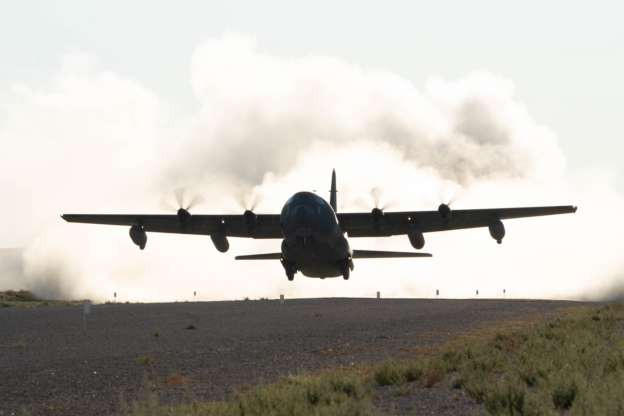 A military cargo aircraft takes off from a dusty dirt air strip.