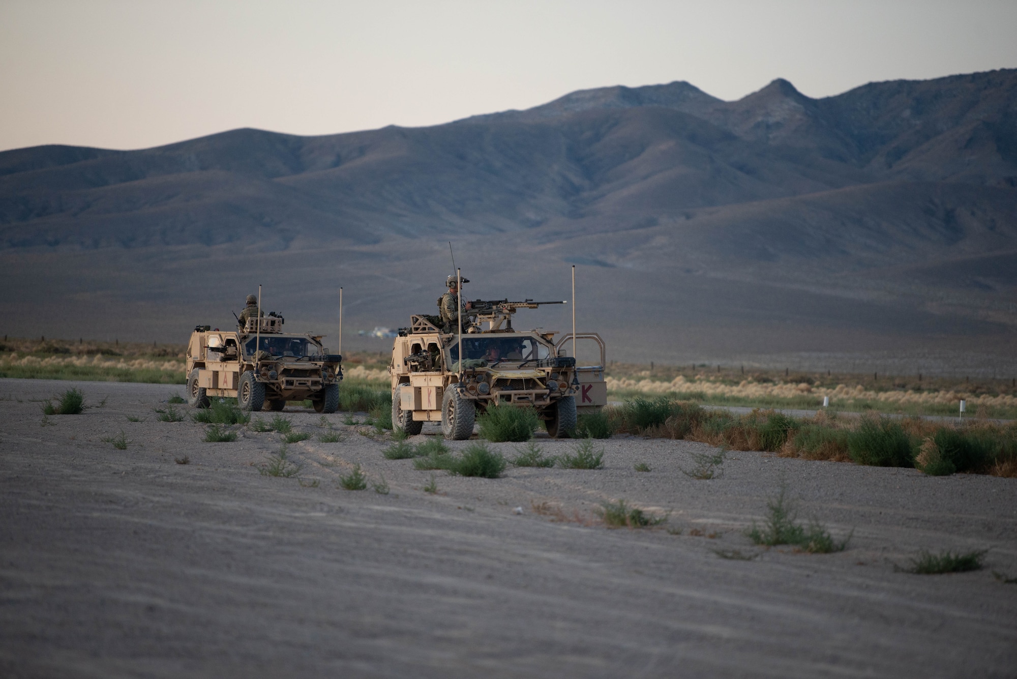 Two military vehicles with mounted heavy weapons idle in a desert field.