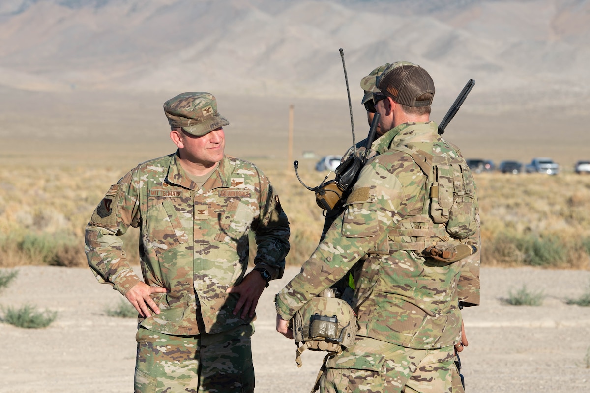 Three men in military uniform stand in a desert field talking.