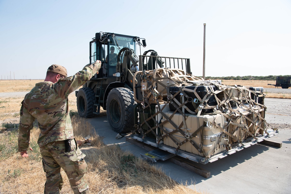 A man in military uniform guides another man driving a piece of heavy equipment as it moves a pallet of equipment.