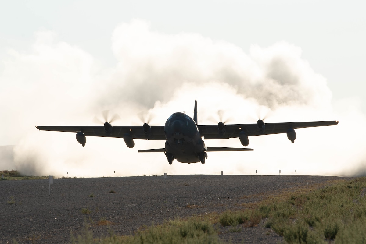 A military cargo aircraft takes off from a dusty dirt air strip.