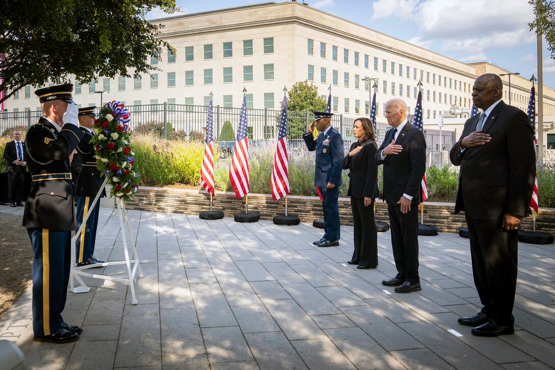 Four leaders stand with hands over heart and one saluting a wreath flanked by two service members.