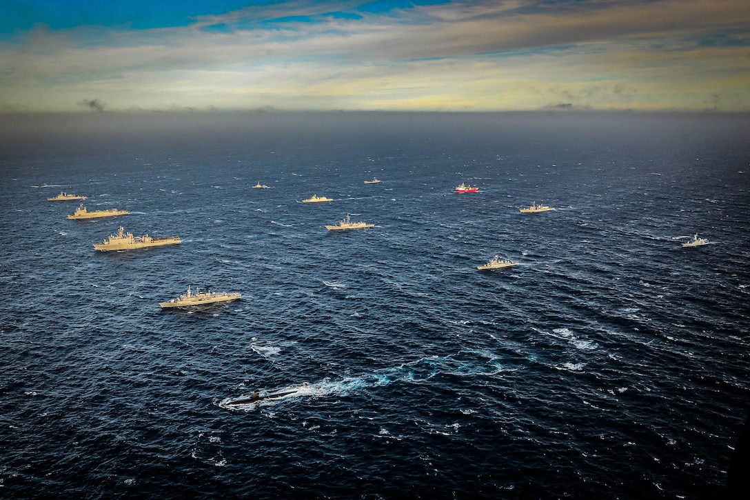 A large group of ships and a submarine steam across a dark blue ocean on a partially cloudy day with a hazy horizon.