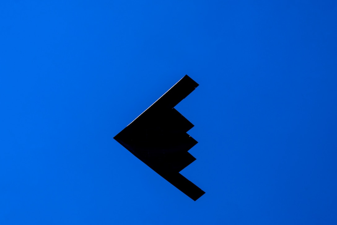 A black B-2 bomber flies across a deep blue sky.