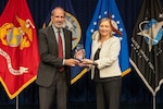 Two people standing and holding an award together in front of four flags.